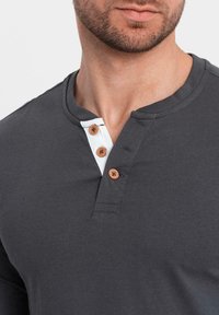 Donkergrijze lange mouwen shirt met een witte contrasterende knoopsluiting en drie bruine knopen. Zacht katoen stof met een Henley ontwerp.
