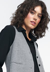 Szary kamizelka w kratę houndstooth z czarną koszulą z kołnierzykiem pod spodem, z fakturowanym materiałem, widocznymi guzikami i detalem kieszeni po lewej stronie.