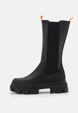 Zwarte laars tot halverwege de kuit met een dikke, grof profielzool, elastische zijpanelen, een bovenwerk van glad leer en oranje aantreklussen aan de voorkant en achterkant.