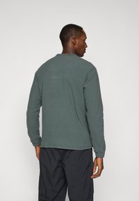 Langärmliges, rundhalsiges Shirt aus dunkelgrünem Stoff. Hat eine lockere Passform mit genähten Säumen und gerippten Bündchen. Keine auffälligen Muster oder Grafiken.