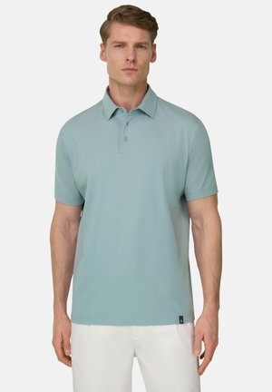 Mand iført lyseblå kortærmet polo og hvide bukser, stående med afslappede arme mod en ensfarvet hvid baggrund.