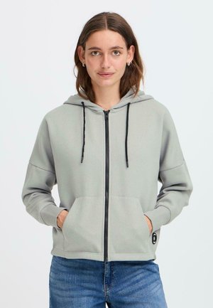 A young woman with shoulder-length hair wears a light grey zip-up hoodie with a hood and front pockets, standing against a plain background.