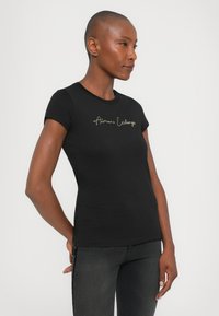 Zwarte katoenen t-shirt met een aansluitend ontwerp en korte mouwen. Bevat een gouden cursieve "Armani Exchange" logo op de borst.