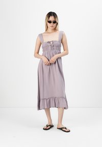 Ljuslila, ärmlös klänning med en smockad bodice, rosettdetalj och volangkant. Bärs med svarta flip-flops och solglasögon.