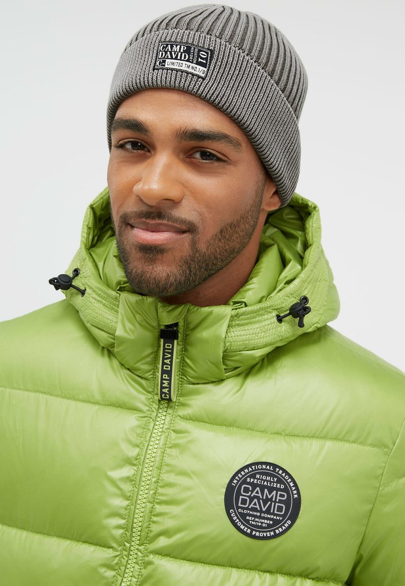 Green puffer jacket with a high collar, featuring a round CAMP DAVID logo badge and a black zipper. Gray ribbed beanie on the head.