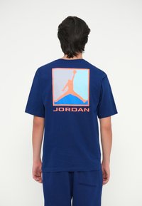 Tumši zilā kokvilnas T-krekls ar lielu oranžu un zilu grafiku, kurā attēlots lecošs figūra, un uzrakstu "JORDAN" mugurā. Īsas piedurknes.