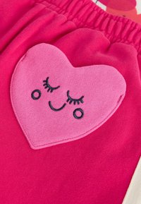 Pantaloni scurți roz din bumbac, cu o pată mare în formă de inimă roz, brodată cu o față zâmbitoare și gene, completată de o talie elastică.