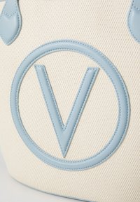 Plátěná taška s logem z světle modré kůže, texturovaným bílým pozadím, kruhovým designem a jemnými detaily šití.