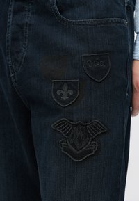 Sötétfarmernadrág három fekete hímzett folttal, pajzs- és szárnymintákkal a jobb combon. Varrt zsebkivágás látható.