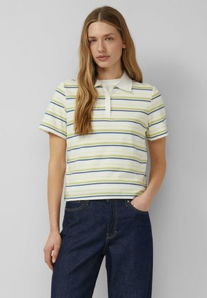 Frau mit langen blonden Haaren, die ein weißes Polohemd mit kurzen Ärmeln und dünnen grünen und blauen horizontalen Streifen sowie dunkelblaue Jeans trägt, eine Hand in der Tasche.