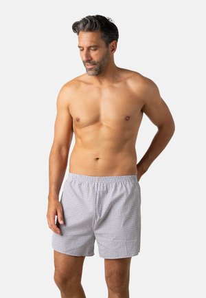 Homme torse nu regardant vers le bas, portant un caleçon à carreaux clairs, debout devant un fond uni gris clair.