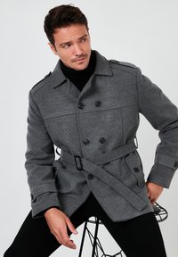 Peacoat grigio in misto lana con bottoni doppi, cintura regolabile, spalline e tasche laterali, indossato su un dolcevita nero.