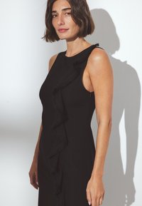 Mustapuku, jossa on hihaton muotoilu ja vasemmalla puolella oleva röyhelökoriste. Sujuva kankaan rakenne, istuva leikkaus ja vetoketjusulku.