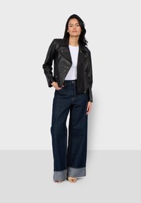 Veste en cuir noir de style motard avec un col cranté, associée à un jean en denim foncé à jambes larges avec un ourlet retroussé. Le modèle porte un haut blanc.