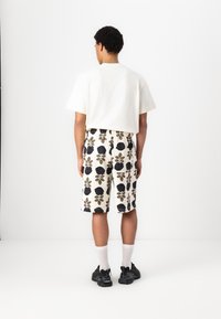 Avslappet passform hvit bomulls t-skjorte kombinert med mønstrede knelengde shorts i krem, svart blomstertrykk og brune detaljer. Svarte joggesko og hvite sokker.