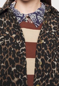 Veste à imprimé léopard en laine noire et brune, superposée sur un pull rayé marron et beige avec un col à motif bleu. Boutons dorés visibles.