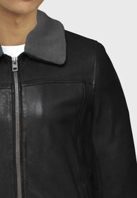 Chaqueta de cuero negra con un cuello de piel gris, que presenta un cierre frontal y costuras detalladas a lo largo de las costuras para añadir textura.