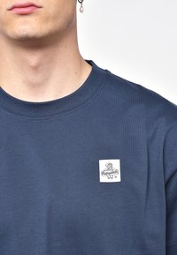 Námořnická modrá bavlněná tričko s kulatým výstřihem a bílou obdélníkovou nášivkou s grafikou psa na levé části hrudi.