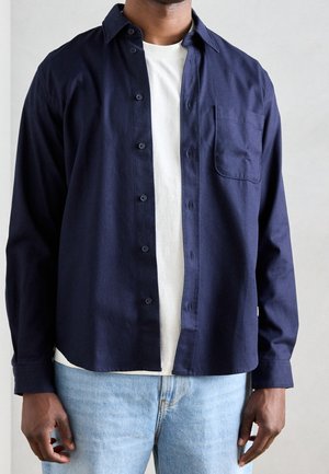 Homme portant une chemise bleu marine déboutonnée sur un t-shirt blanc et un jean bleu clair sur fond uni.