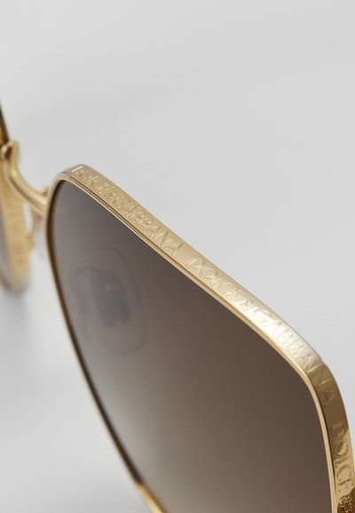 Goldene Metall-Sonnenbrille mit braunen Gläsern, ausgestattet mit gravierten Logo-Details am Rahmen. Winkelige Form mit glänzender Oberfläche und strukturierten Akzenten.