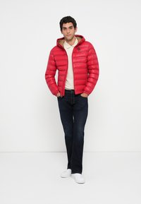 Man standing with hands in pockets wearing red puffer jacket, cream polo shirt, dark jeans, and white sneakers on white background.