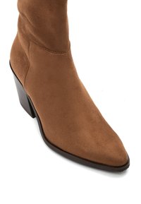 Stivale alla caviglia in suede marrone con punta a mandorla e tacco a blocco impilato su uno sfondo bianco.