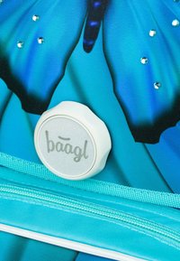 Modrá taška má design motýla, třpytivé kamínky a kulatý bílý knoflík s logem "baagl." Obsahuje texturovaný zipový akcent.
