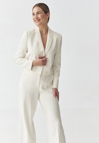 Bílý přizpůsobený blazer s klopami, v kombinaci s odpovídajícími kalhotami s vysokým pasem. Hladká tkanina, čisté linie a přiléhavý silueta.