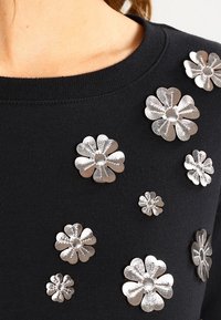 Svart tröja med metalliska silverblommor i varierande storlekar som bildar en strukturerad design på framsidan.