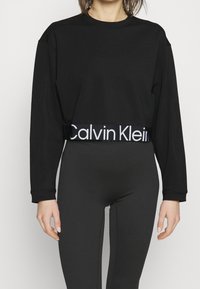 Svart croppad sweatshirt med långa ärmar, med en kontrasterande vit Calvin Klein-logoband i nederkant och en slät tygtextur.
