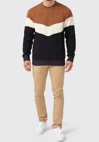 Pull tricoté avec un motif chevron en marron, crème et marine, avec des poignets et un ourlet côtelés. Porté avec un pantalon beige et des baskets blanches.