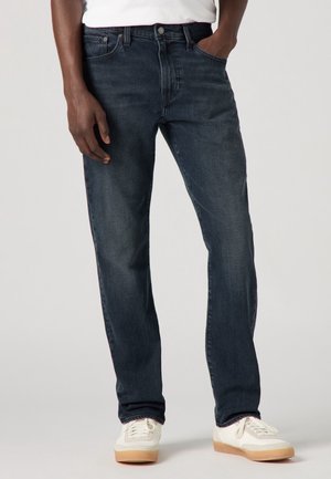 Homme portant un jean droit bleu foncé et des baskets blanches, une main dans la poche et l'autre détendue à ses côtés.