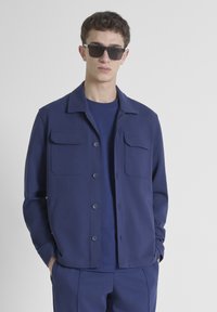 Chaqueta azul marino de tela ligera con cuello, cierre de botones y dos bolsillos en el pecho. Llevada sobre una camisa a juego, combinada con gafas de sol.