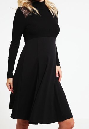Vrouw draagt een zwarte jurk met lange mouwen, kanten schouderdetails en een uitlopende rok tegen een witte achtergrond.