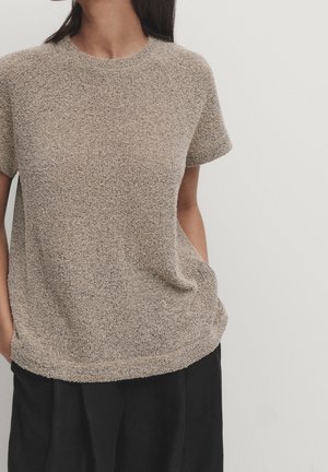 Femme portant un haut beige texturé à manches courtes avec un pantalon noir, mains dans les poches, debout devant un fond clair uni.