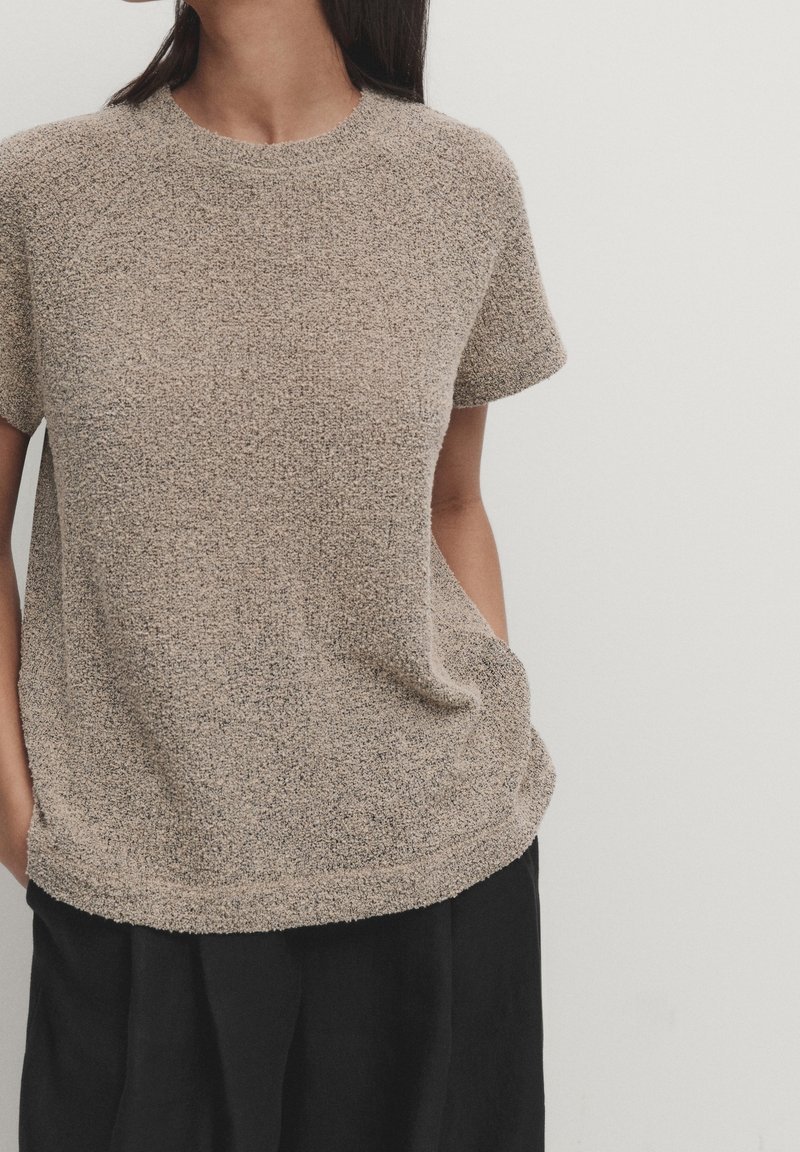 Femme portant un haut beige texturé à manches courtes avec un pantalon noir, mains dans les poches, debout devant un fond clair uni.