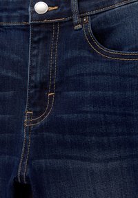 Dunkelblaue Denim-Jeans Nahaufnahme mit Vordertasche, gelber Naht und silbernem Knopf mit Markentext.
