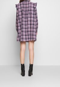 Vestido camisero de cuadros en púrpura, negro y rosa con detalles de volantes en los hombros. Combinado con botas de tacón negro hasta el tobillo.