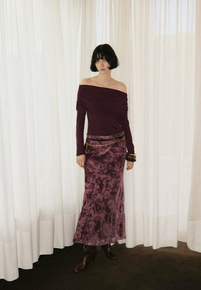 Haut bordeaux à épaules dénudées avec des manches drapées, associé à une longue jupe plissée prune. Les accessoires comprennent des bracelets noir et or, ainsi que des bottes à talons marron.