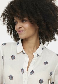 Chemise blanche à boutons en tissu léger avec des motifs géométriques marron et bleu brodés de manière complexe.