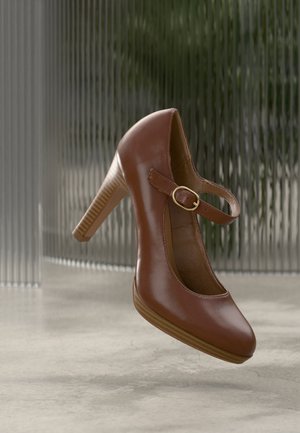 Chaussure Mary Jane à talon haut en cuir marron avec bout arrondi et boucle dorée, présentée sur une surface réfléchissante neutre et un fond en verre texturé.