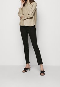 Kvinna iklädd en beige långärmad skjorta med knäppning, svarta slim fit-byxor med fransade fållar och svarta klacksandaler med knutdetalj, stående framför en vit bakgrund.