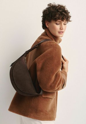 Brązowa zamszowa torba na ramię o zaokrąglonym kształcie, solidnym pasku i zamku błyskawicznym, noszona na brązowej teksturowanej kurtce z polaru.