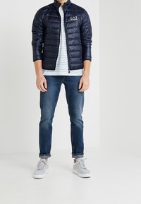 Námořnická puffer bunda s lesklým povrchem, zapínáním na zip a logem. Nositá přes pruhované tričko a v kombinaci s modrými dž�íny a šedými teniskami.