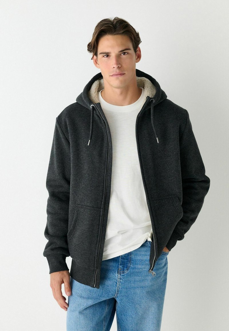 Dunkelgrauer Reißverschluss-Hoodie mit einem fleecegefütterten Kapuzen, Fronttaschen und Zugbändern, kombiniert mit einem weißen Shirt und blauen Jeans.