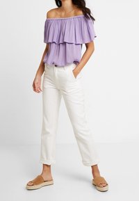 Carmen-Bluse in Flieder mit lagenweisen Rüschen, kombiniert mit weißen, hochgekrempelten Hosen und beigen Rutsch-Sandalen mit Jutesohle.