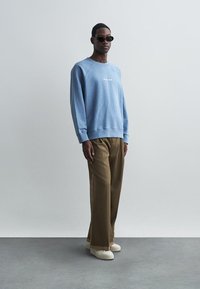 Lyseblå genser med ribbede mansjetter, med hvit tekst på brystet. Brune vidbenede bukser og beige slip-on-sko fullfører antrekket.