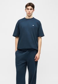 Νέος άνδρας στέκεται στραμμένος προς τα εμπρός, φορώντας μια μπλούζα σε σκούρο μπλε oversized και χαλαρό παντελόνι σε ταιριαστό χρώμα, μπροστά σε απλό φωτεινό φόντο.