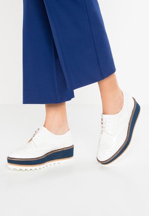 Person iført blå cropped bukser og hvide platform brogue sko med marineblå og korksåler, stående på en hvid gulv.