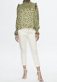 Blusa verde con motivo floreale, colletto alto arricciato e maniche con volant, abbinata a pantaloni crema alla caviglia e sandali con cinturini.